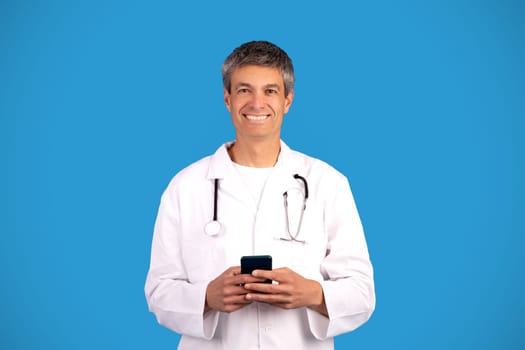 Happy physician doctor man holding smartphone standing on blue background