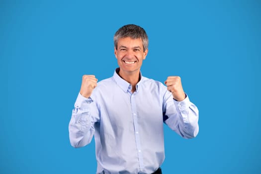 Mature businessman shaking fists celebrating luck over blue background