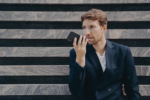 Male executive worker dressed formally holds smartphone near mouth talks on speakerphone