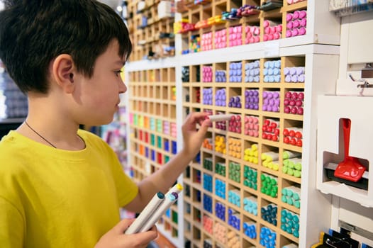 Adorable teen boy, school kid takes out watercolor markers from the shelf, choosing drawing and sketching tools while shopping in an office supply shop. School stationery store. Creative Art shop