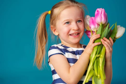 Smiling little blonde girl with tulips bouquet against blue background