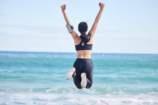 Happy woman, fitness and jump in achievement on beach for winning, workout success or outdoor exercise. Rear view of excited female person in celebration for accomplishment or training by the ocean