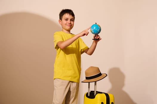 Smiling teen boy in yellow t-shirt, pointing at a travel destination on the globe, travelling abroad for weekend getaway