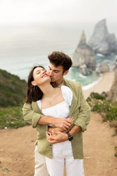 Lovely young european couple enjoying date on coastline, man embracing lady from back and kissing her, standing on nature with perfect view at ocean shore, vertical shot