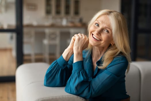 Portrait of happy caucasian mature woman resting on sofa at home interior, looking and smiling at camera, copy space