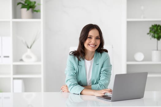 Cheerful young caucasian business woman in suit with computer enjoys professional occupation in office
