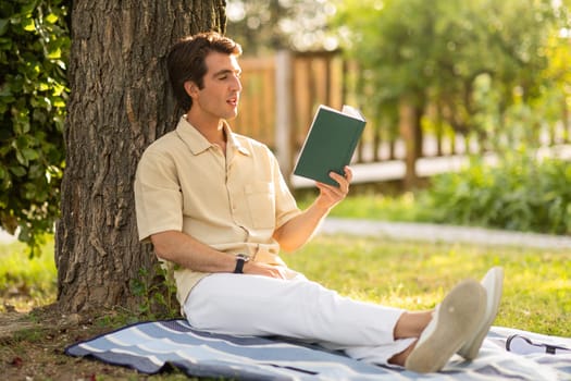 Carefree young guy sitting under tree at garden, reading book