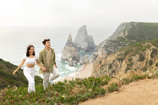 Happy european couple in love holding hands while walking on nature with beautiful view on ocean shore, having date