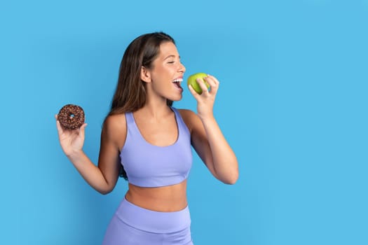 Cheerful slim young caucasian lady in sportswear with donut in hand bites green apple