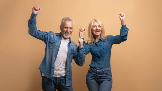 Emotional thrilled happy senior people man and woman friends or couple raising hands up and exclaiming on beige background. Cheerful elderly family spouses celebrating success