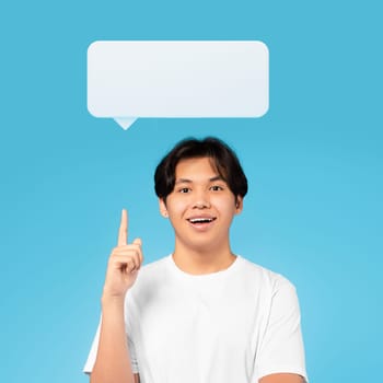 Your opinion matters concept. Excited handsome millennial asian guy in white t-shirt pointing index finger up at white blank communication bubble, smiling on blue studio background, having nice idea