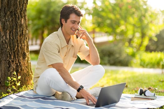 Cheerful young man digital nomad resting at park, using gadgets