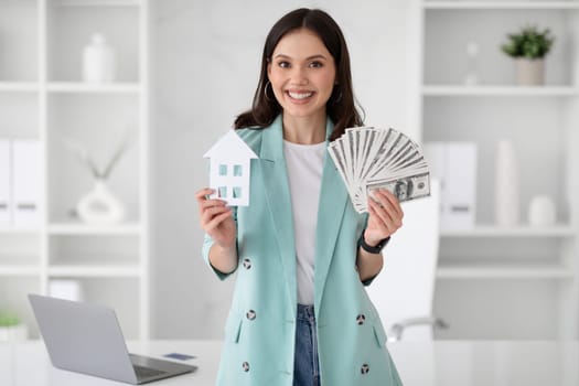 Cheerful young caucasian businesswoman realtor in suit hold a lot of money, dollars and house