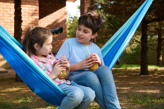 Close-up Caucasian adorable school kids drinking smoothie while relaxing on hammock in the backyard. Children. Leisure. Lifestyle
