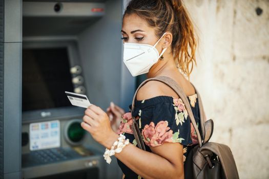 Side view of a young woman is wearing N95 protective mask while taking money from ATM machine during Covid-19 pandemic.