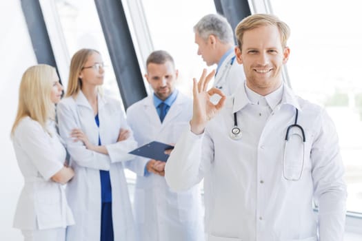 Team of medical professionals, young doctor looking at camera, smiling, showing ok sign