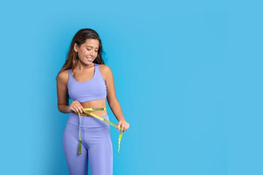 Positive millennial european lady in sportswear with measuring tape measures waist