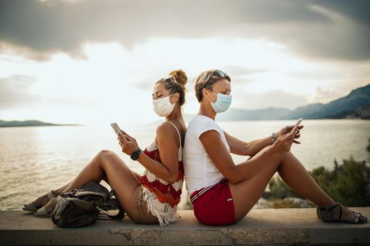 Shot of a happy female friends with protective mask spending time on seaside and surfing social media during exploring a Mediterranean at corona pandemic. 