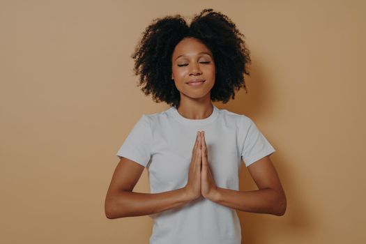 African american young woman holding hands clasped together and praying on beige background