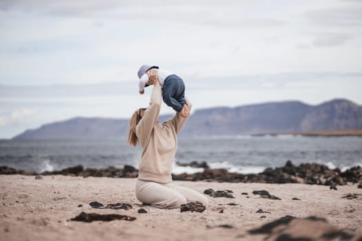 Mother enjoying winter vacations holding, playing and lifting his infant baby boy son high in the air on sandy beach on Lanzarote island, Spain. Family travel and vacations concept
