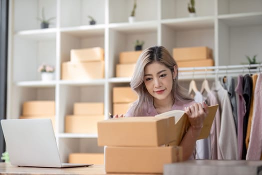 Female entrepreneur writes on parcel box after reading address on notebook. Online seller concept