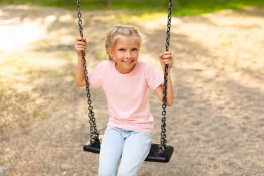 Happy schoolgirl sitting on swing while walking on playground, girl looking and smiling at camera, copy space