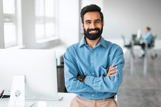 Successful young indian businessman in shirt standing with folded arms near desk, smiling at camera, office interior