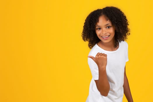 Positive teenage black pupil girl in white t-shirt point finger on empty space, recommends study