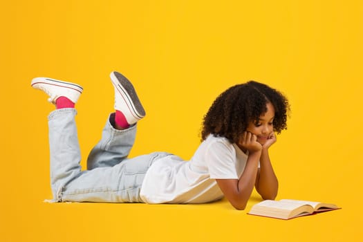 Smiling teenage black pupil girl in white t-shirt reads book, lies on floor, isolated on yellow background