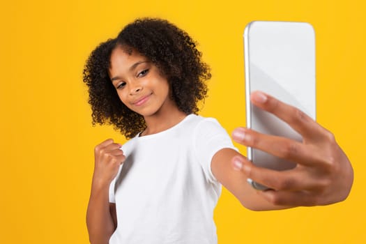 Strong glad teenage black pupil girl in white t-shirt takes selfie on phone, makes success, victory sign with hand