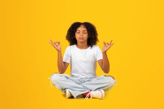 Positive teenage black pupil girl with closed eyes, sit on floor, meditating, isolated on yellow background