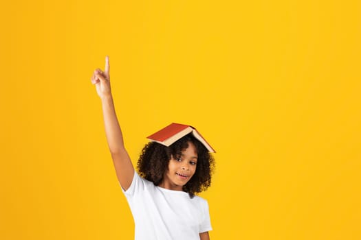 Glad teenage black pupil girl in white t-shirt with book on head, raises hand up, has fun