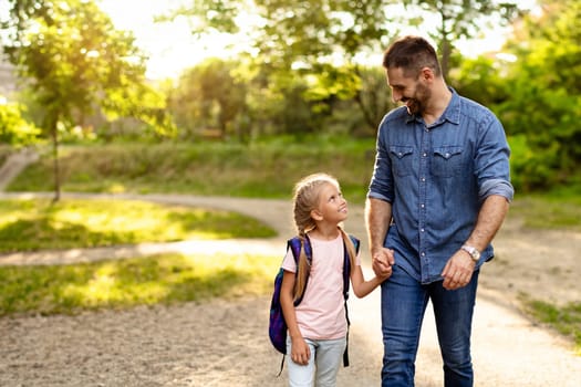 Happy middle aged father holding hand with smart schoolgirl and communicating while walking on path in park, free space