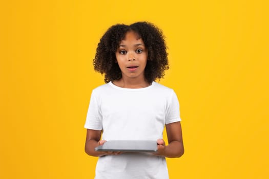 Shocked positive teenage black pupil girl in white t-shirt use tablet, isolated on yellow background, studio