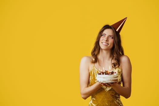 Satisfied dreamy woman in hat making wish and hold cake with candles, looking at free space, yellow background