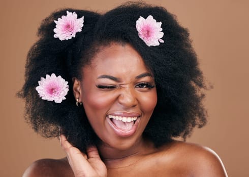 Flowers, face wink and black woman in afro hair care, excited and beauty in studio isolated on a brown background. Portrait, floral hairstyle cosmetic and natural African model flirt in organic salon