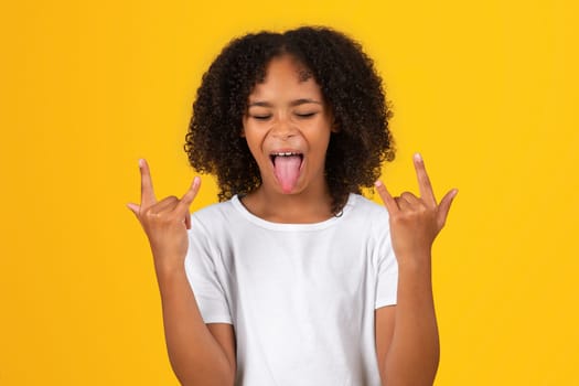 Glad excited teenage black pupil girl in white t-shirt show tongue, make goat gesture with hands
