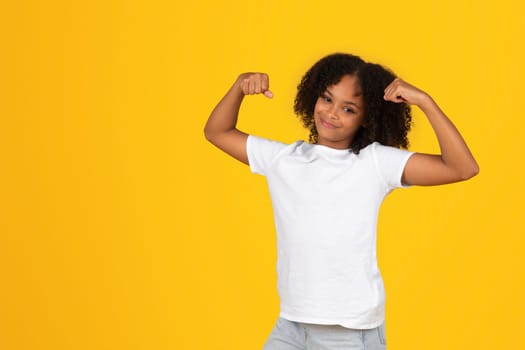 Positive teenage black pupil girl in white t-shirt shows biceps muscles
