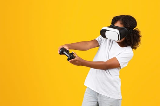Positive teenage black pupil girl in white t-shirt, VR glasses with joystick enjoys spare time, playing online game
