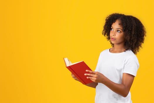 Positive surprised teenage black pupil girl in white t-shirt reads book, looks at empty space