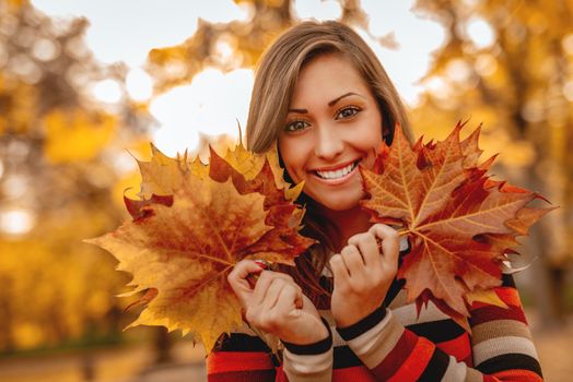 Cute young woman enjoying in sunny forest in autumn colors. She is holding many leaves and looking at camera behind them.