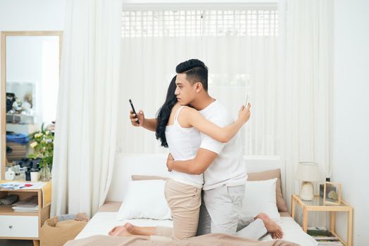 Young couple in the bedroom. Side view of unfaithful man and woman are cheating on the phone while hugging each other