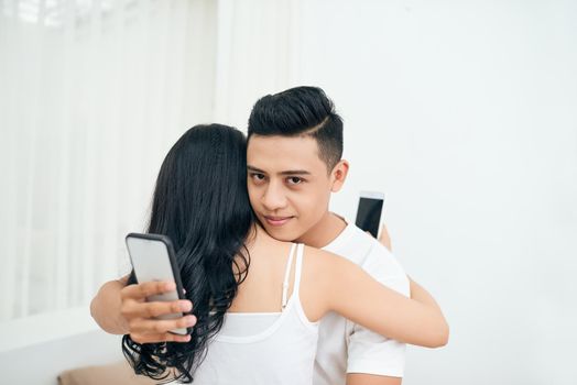 Young couple in the bedroom. Smiling unfaithful man is cheating and texting lover on the phone while hugging his girlfriend