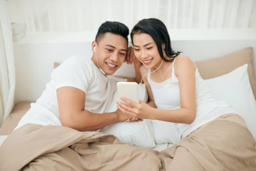 Indoor photo of loving couple lying on white sheets with smartphone.