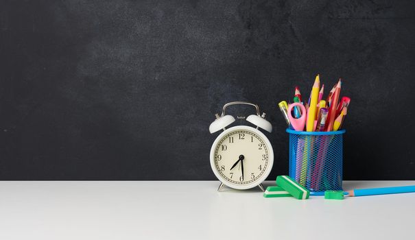 white round alarm clock and a metal glass with pens, pencils and felt-tip pens on the background of an empty black chalk board