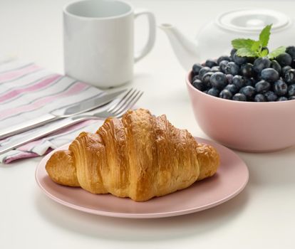 Baked crisp croissant on a wooden board, a plate with blueberry, a white ceramic brew and a cup on a white table. Breakfast