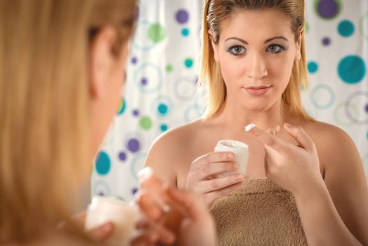 Beautiful young woman applying cream on her face in front a mirror in the bathroom at home. 