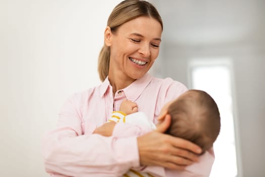 Loving smiling mother lulling her baby daughter to sleep, carrying her in arms, standing in bedroom interior at home