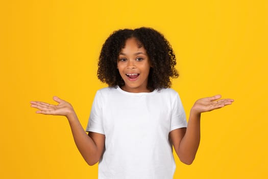 Glad shocked adolescent black girl in white t-shirt with open mouth spreads arms to sides, holds empty space