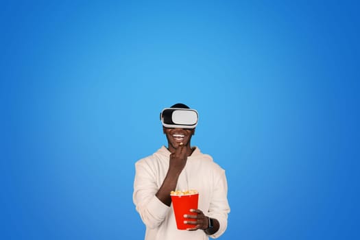 Cheerful young african american guy in hoodie and vr glasses watch video, eating popcorn